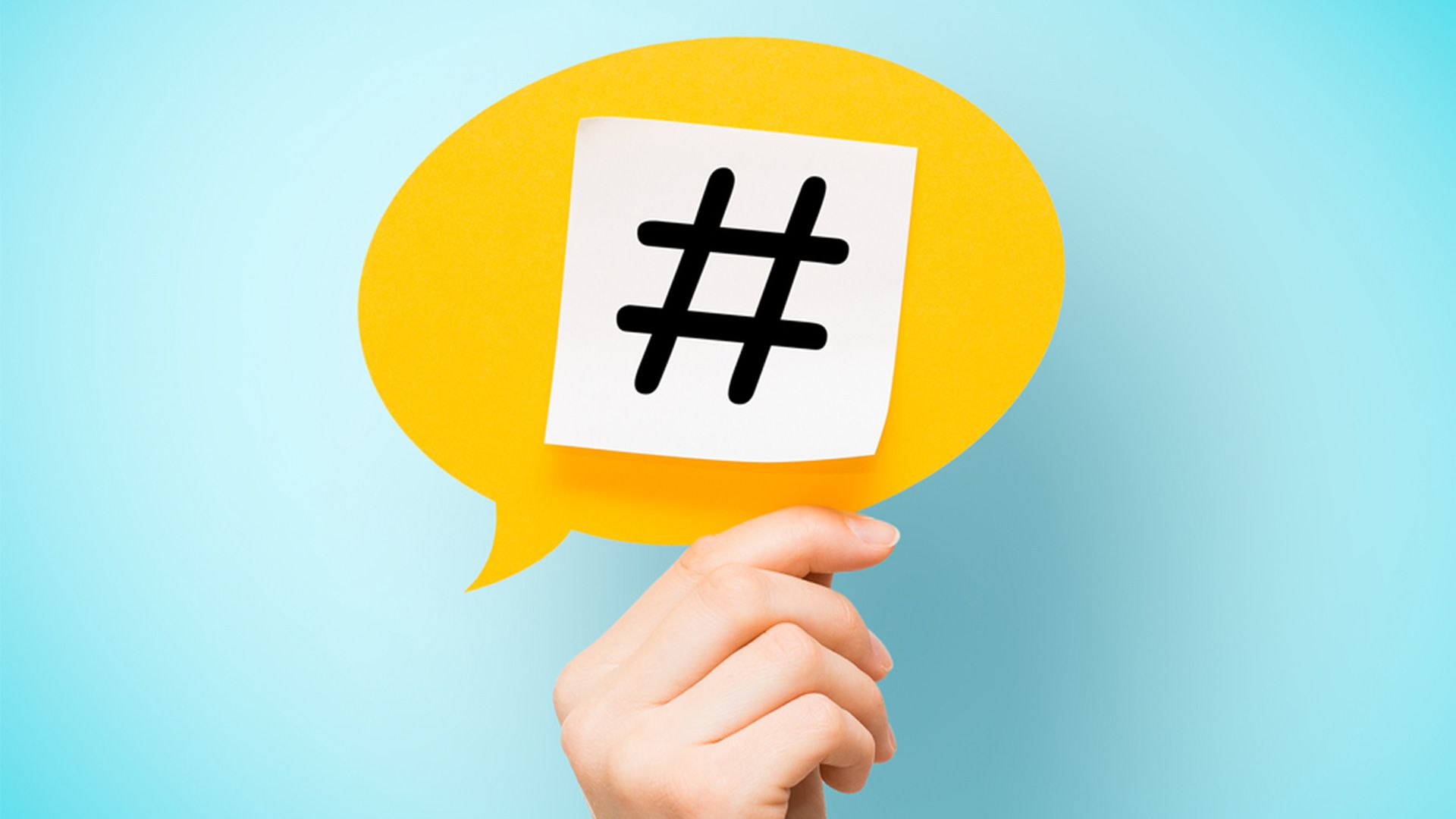 Eine Hand hält eine gelbe Sprechblase mit Hashtag darauf vor hellblauem Hintergrund
