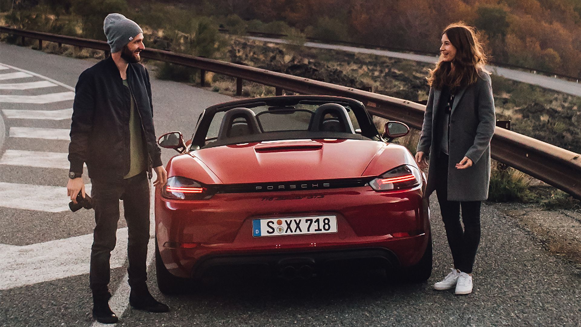 Ein Mann und eine Fau stehen sich lächelnd gegenüber. Zwischen ihnen steht ein rotes Porsche Cabriolet.
