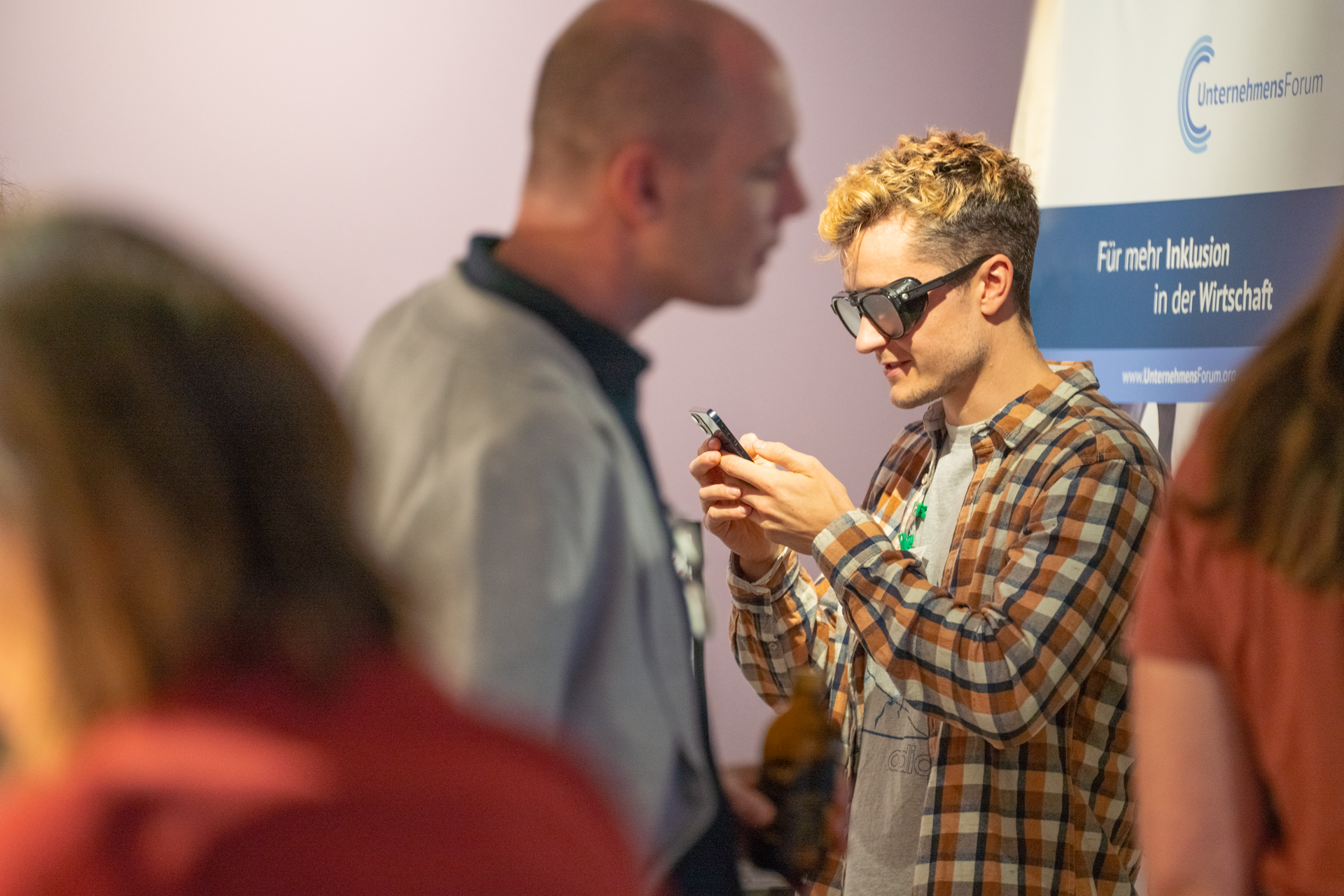 Ein Teilnehmer der UDG Executive Lounge "Accessibility Special" mit und bei Google testet eine markante schwarze Brille, die Menschen mit Sehbehinderung die Nutzung von Smartphones erleichtert. Im Vordergrund sind unscharf weitere Teilnehmende der Lounge zu sehen.