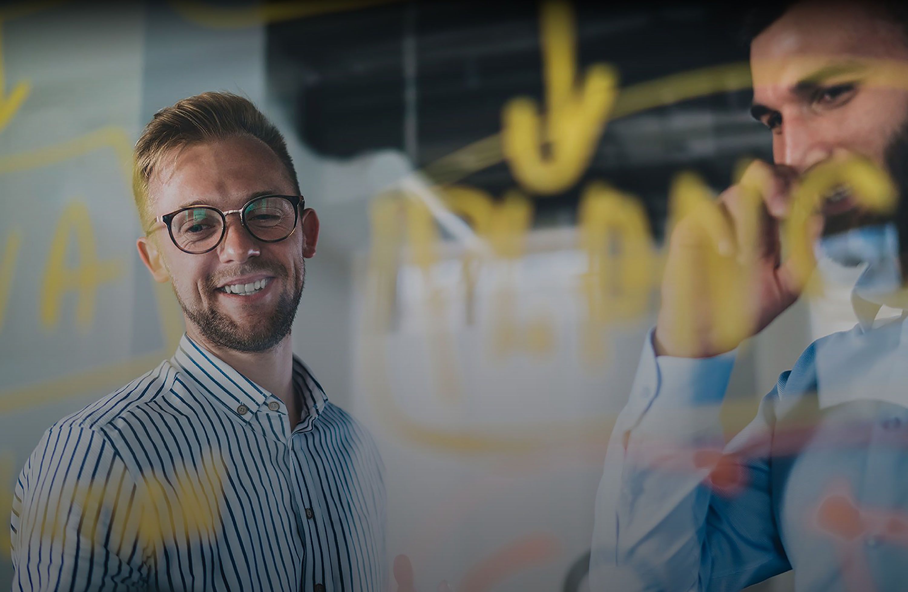 Two men in an office, one smiling and wearing glasses, the other thinking. They are behind a glass wall with yellow writing.