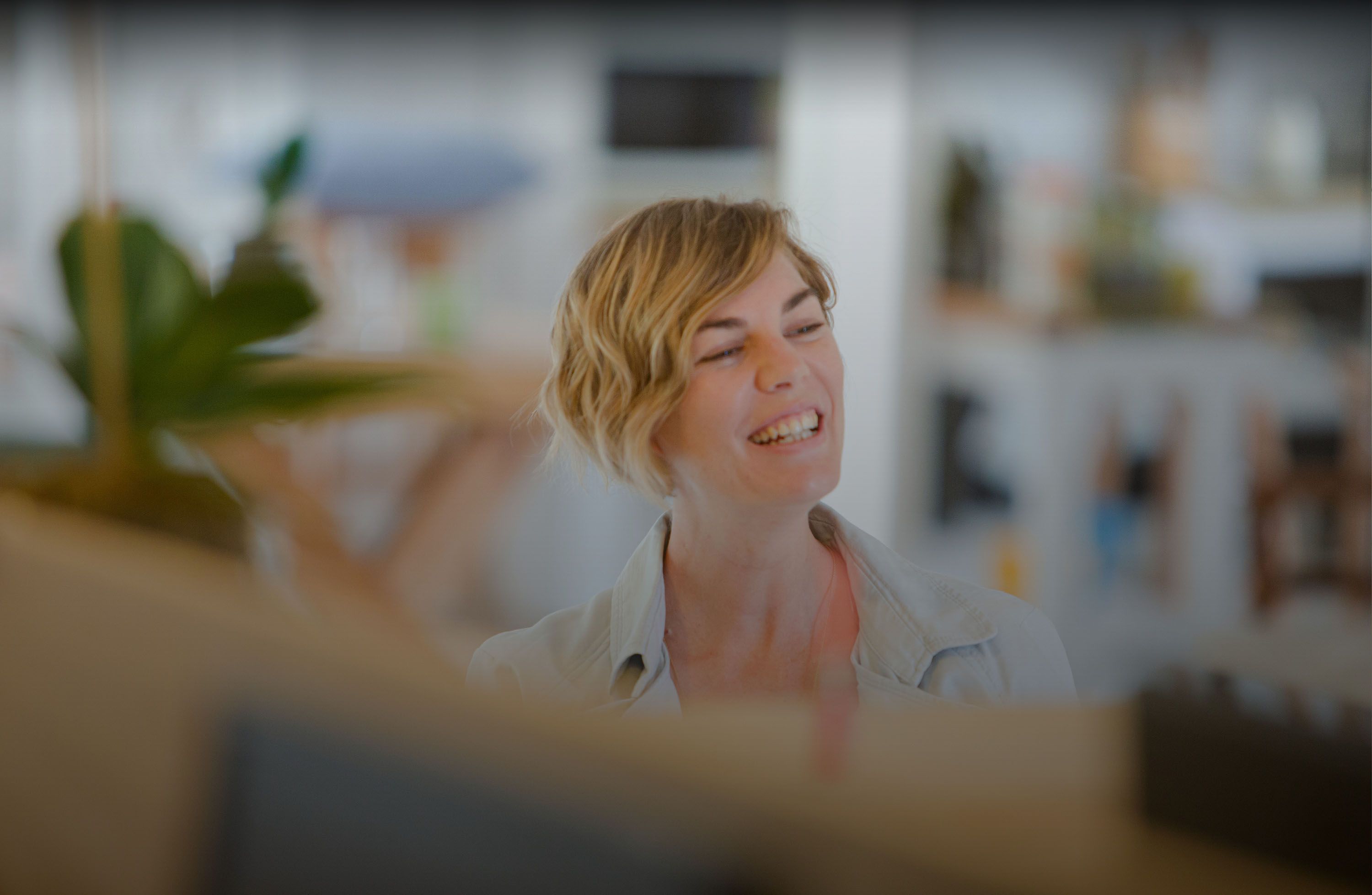 Eine Frau mit kurzen blonden Haaren in einer hellen, unscharfen Innenraumkulisse mit Pflanzen im Vordergrund.