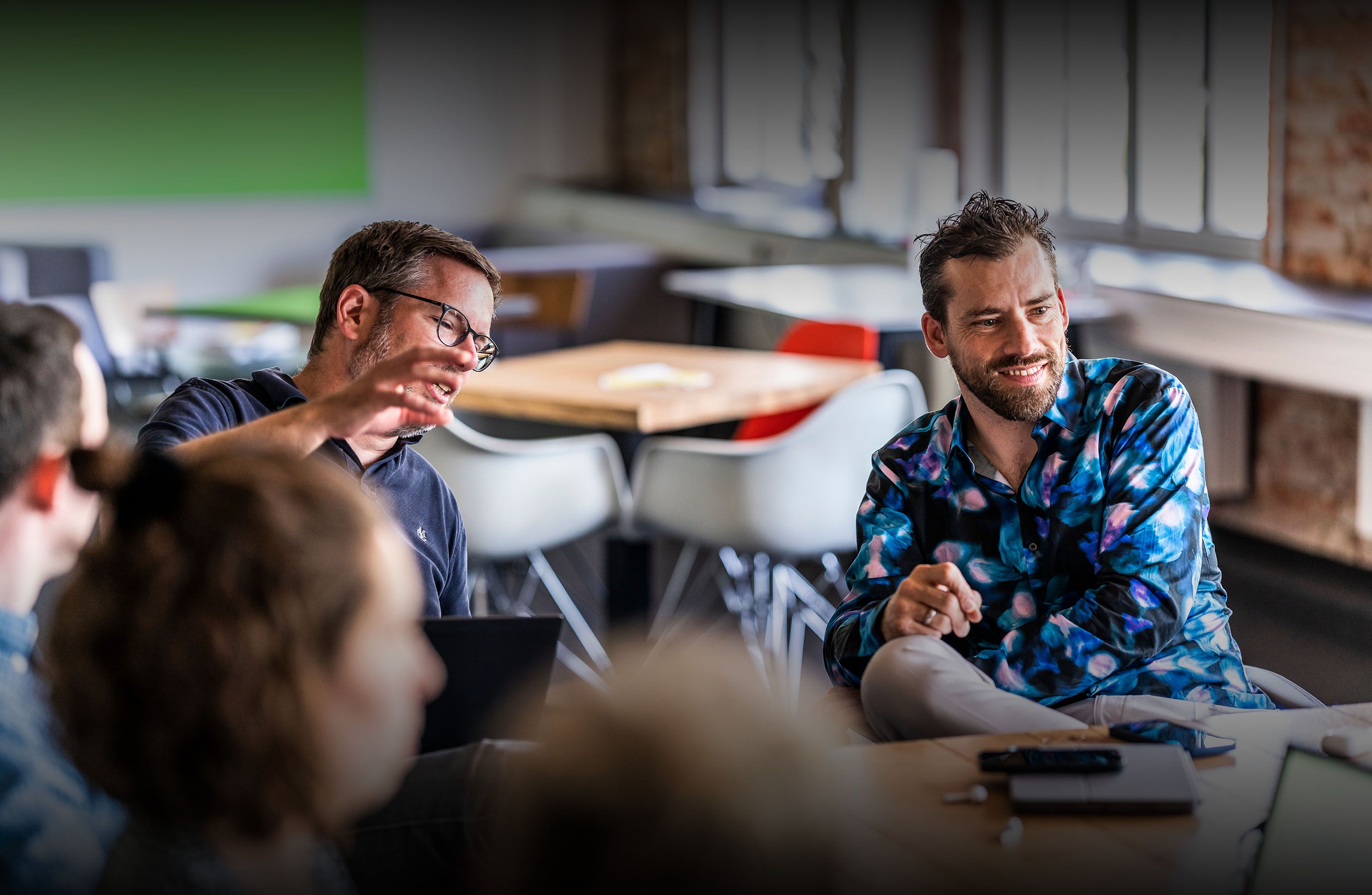 Zwei Männer unterhalten sich an einem Tisch in einem hellen, modernen Raum. Der eine gestikuliert lebhaft, der andere hört mit einem Lächeln zu.