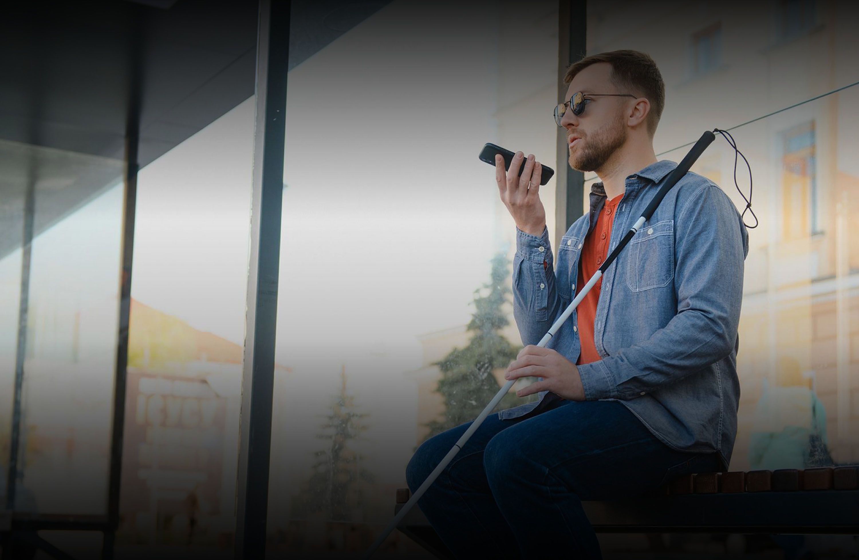 Blinder Mann mit Sonnenbrille und Blindenstab sitzt vor Gebäude auf einer Bank und spricht in sein Mobiltelefon